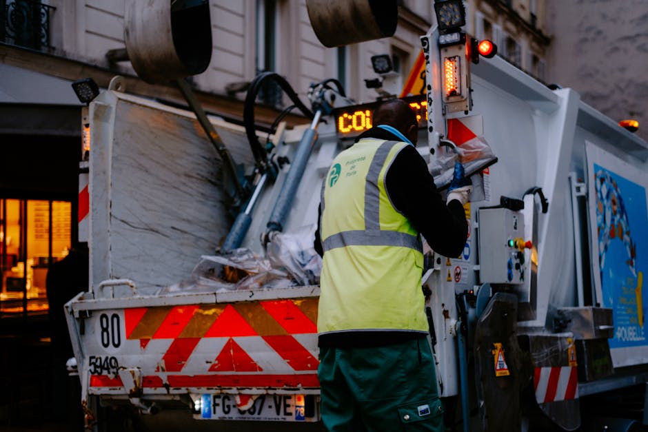 A man dressed in reflective orange trousers, a white high-visibility jacket, and a blue cap is using a leaf blower or similar equipment to clear litter from a grassy park or open area. The equipment h